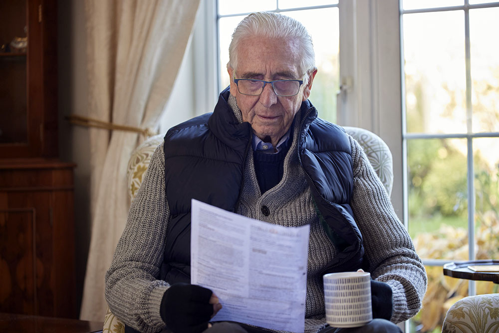 Older man wearing warm clothes, drinking a warm drink and reading a piece of paper