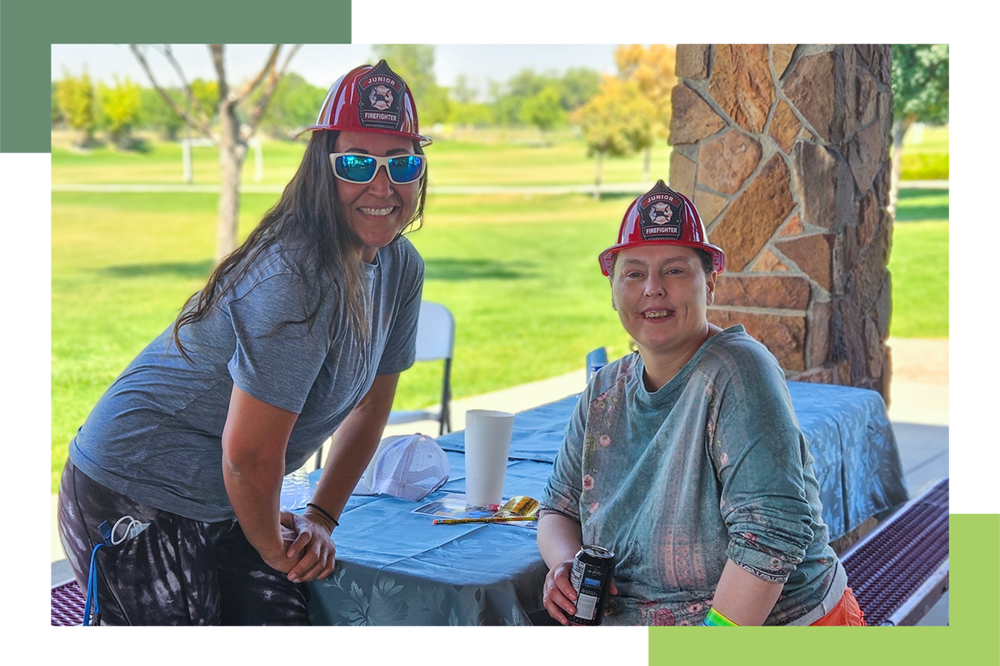 A female individual with IDD wearing a plastic, red firefighter helmet takes a break from eating outdoors for a photo with a female GJRC staff member, who is also wearing a plastic, red firefighter helmet.