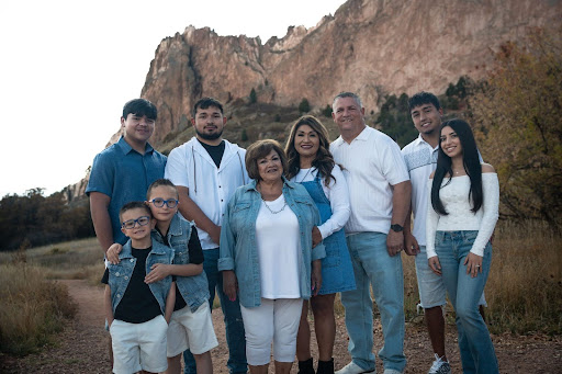 A diverse family of eight people poses for a portrait on a dirt path with a large rocky mountain in the background.