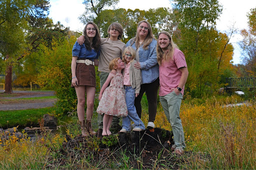 A family of six, including four children and two adults, is posing for a photo outdoors in autumn, standing on a tree stump with colorful foliage and a stream in the background.