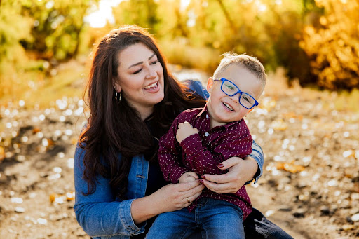 A woman in a denim jacket holds a young boy in blue glasses who is laughing in a sunlit fall outdoor setting.