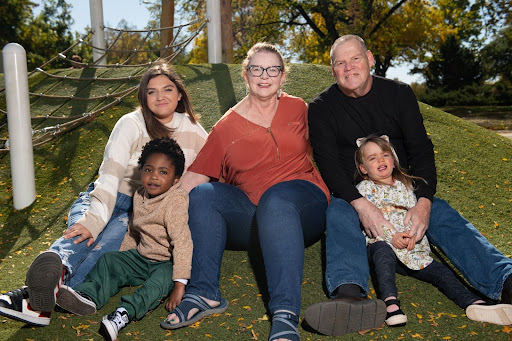 A multiracial family of five, including two adults, two children young children, and an adult child, sits on a grassy hill outdoors.