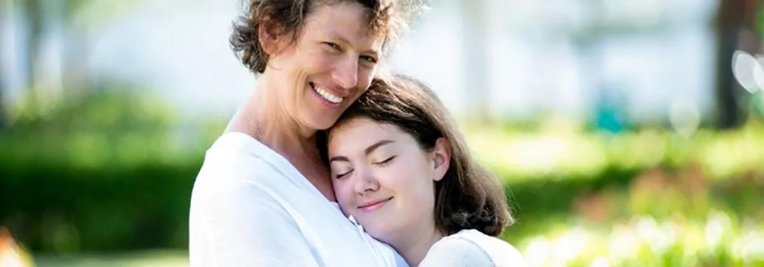 A middle-aged woman with short, curly hair smiles and hugs a teenage girl. The girl, with her eyes closed and a peaceful expression, rests her head on the woman's shoulder.